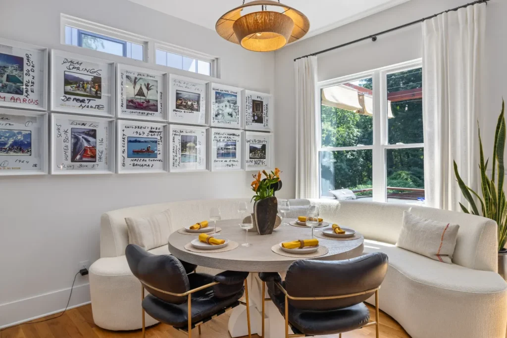 A modern dining nook, ideal for an str photographer, features a round table set for four, white curved banquette seating, black chairs, and large windows. Artworks adorn the wall beneath a contemporary pendant lamp above the table.