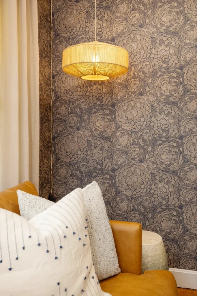 A cozy corner with a tan leather sofa, white and patterned pillows, a round woven pendant light, and a blue floral-patterned wallpapered wall in the background.