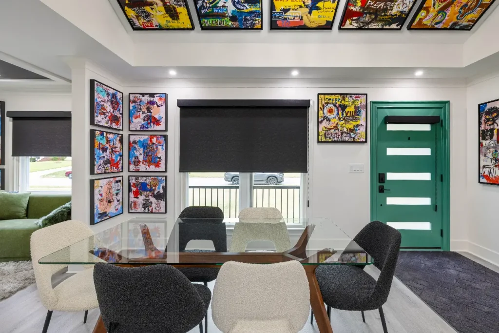Modern dining room with a glass table, six chairs, a green front door, black-out window shades, and colorful comic art displayed on the white walls and ceiling. Natural light streams in through the windows.