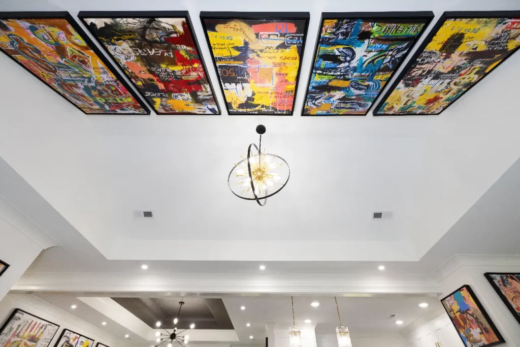 Colorful abstract paintings are displayed on the ceiling of a modern white room with recessed lighting and a central decorative chandelier. More art and lighting fixtures are visible on the walls and ceiling below.