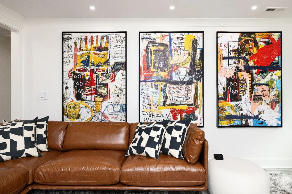 A modern living room with a brown leather sofa, black-and-white patterned pillows, and three large abstract art pieces with bold colors and text hanging on a white wall above the couch.