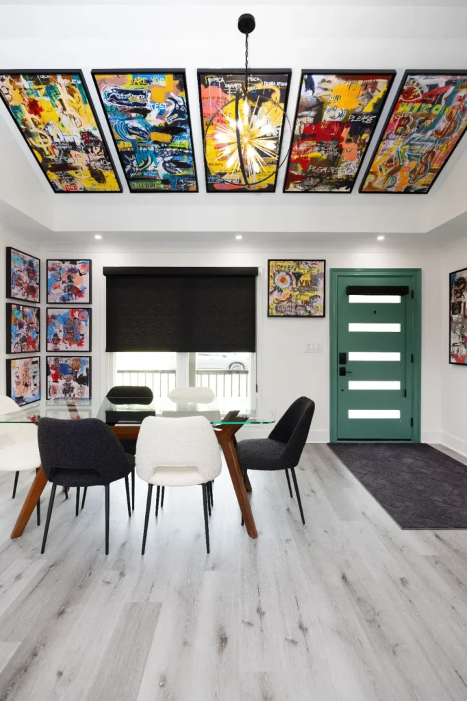 A modern dining room with a glass table, assorted chairs, light wood floor, and a teal front door. Colorful abstract art covers the ceiling and walls, adding vibrant energy to the space.