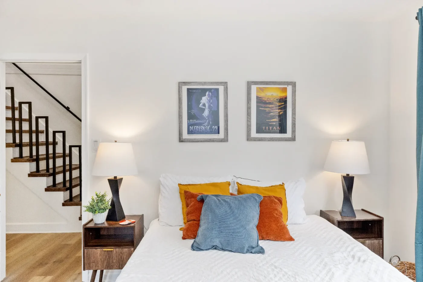 A neatly made bed with white linens, orange and blue pillows, and two bedside tables with lamps. Above the bed are two framed posters. A staircase is visible through an open doorway on the left.