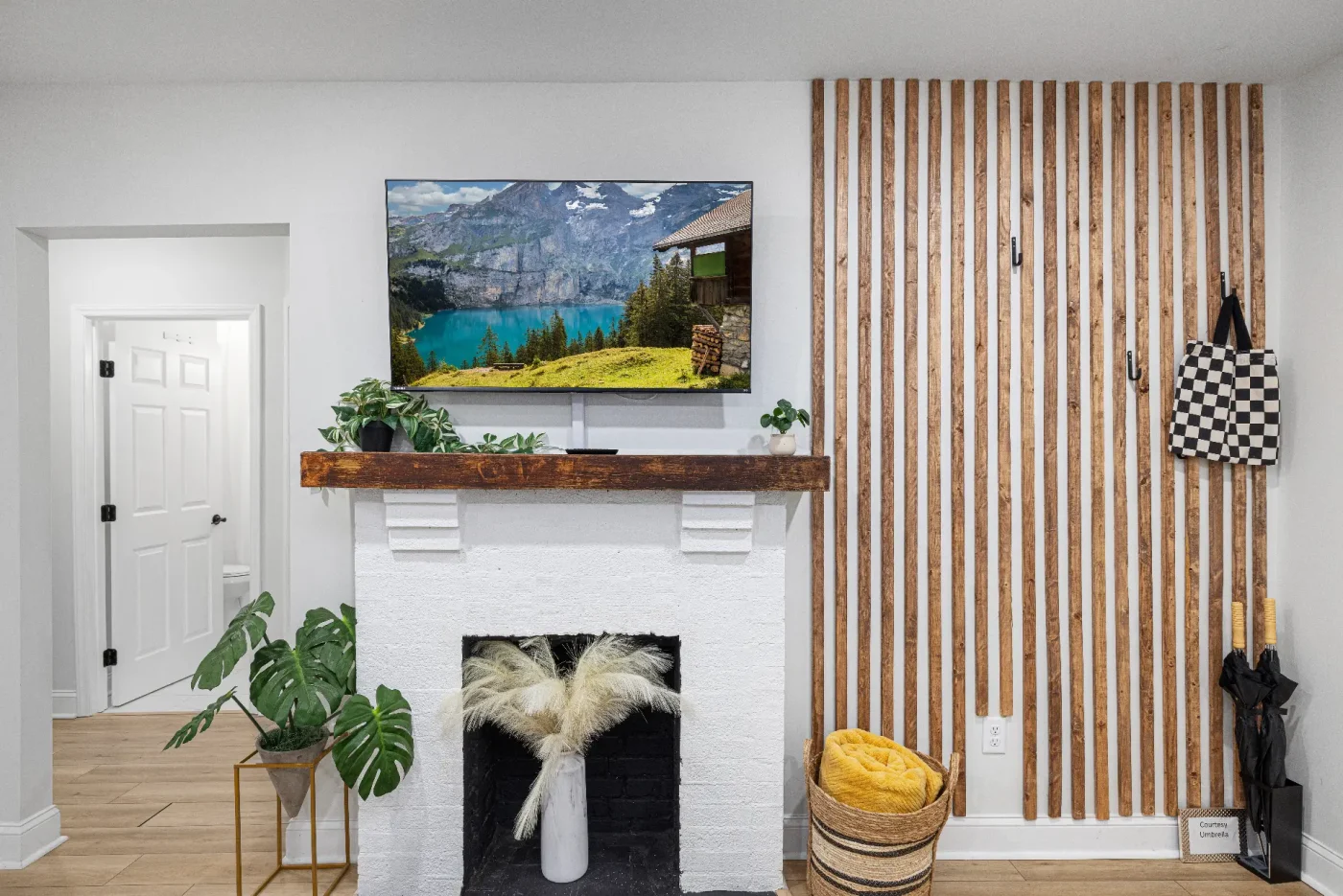 A modern living room features a white fireplace with a wooden mantel, plants, a TV displaying a mountain lake scene, a slatted wood accent wall, a basket with a yellow blanket, and an umbrella by the door.