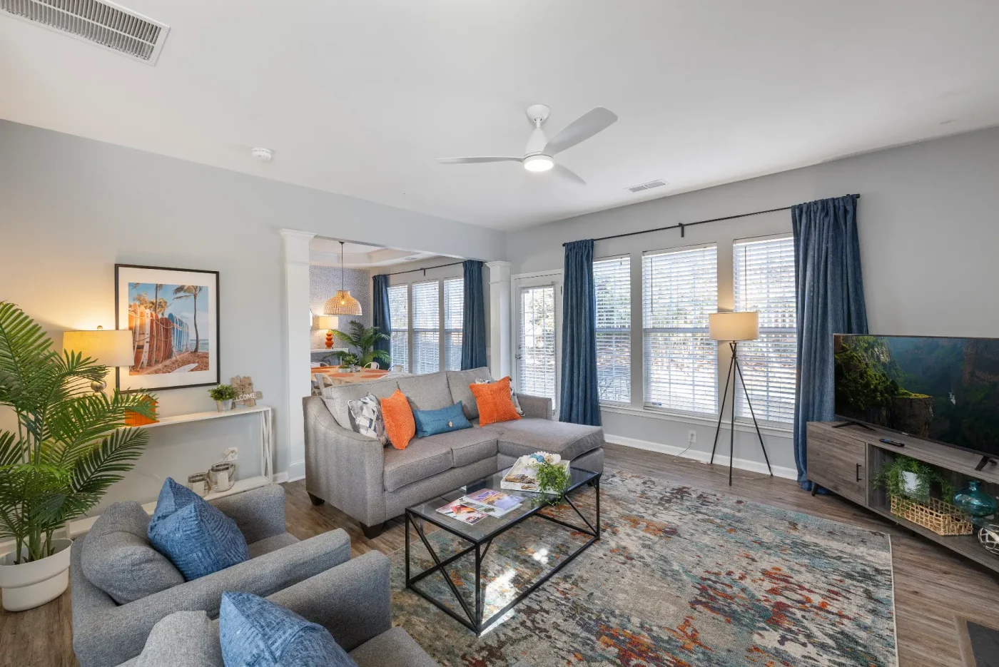 Modern living room with gray sofas, orange and blue pillows, a colorful rug, a coffee table, TV, large windows with blue curtains, potted plants, and wall art. Sunlight brightens the spacious, inviting space.